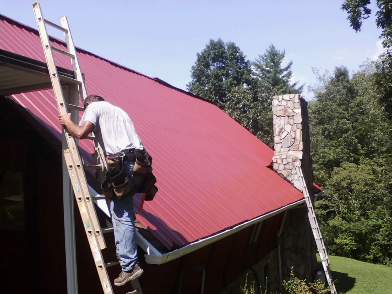 Roofer on a ladder beside a red metal roof and stone chimney — Soffit & Fascia Repair by Roofing Brothers in Ashland